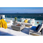 Outdoor patio furniture set on a white-decked terrace overlooking the ocean, including a white sectional sofa with patterned and yellow pillows, a gray fire pit table, a gray armchair with blue and dark pillows, and a yellow throw blanket. The brand name ELEMENTI appears in the lower-left corner.