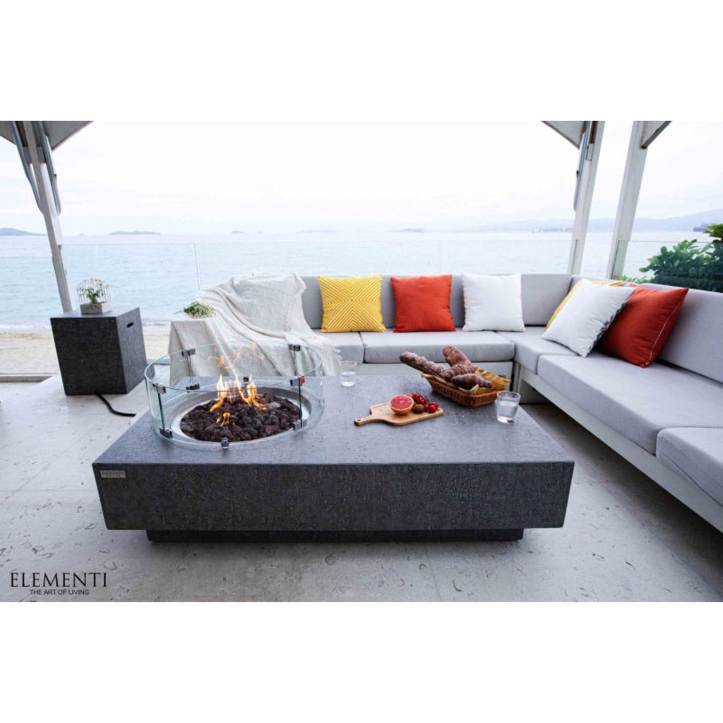 A modern outdoor patio scene with a large rectangular dark gray fire pit table in the foreground, a light gray sectional sofa with yellow, red, and white pillows, and an ocean view in the background. The brand name ELEMENTI appears in the bottom-left corner.