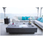A modern outdoor patio scene featuring a large dark gray rectangular coffee table with a circular inset, set in front of a light gray sectional sofa with turquoise pillows. The scene is styled with a three-tiered tea stand, cups, and a small side table, with the ocean visible in the background.