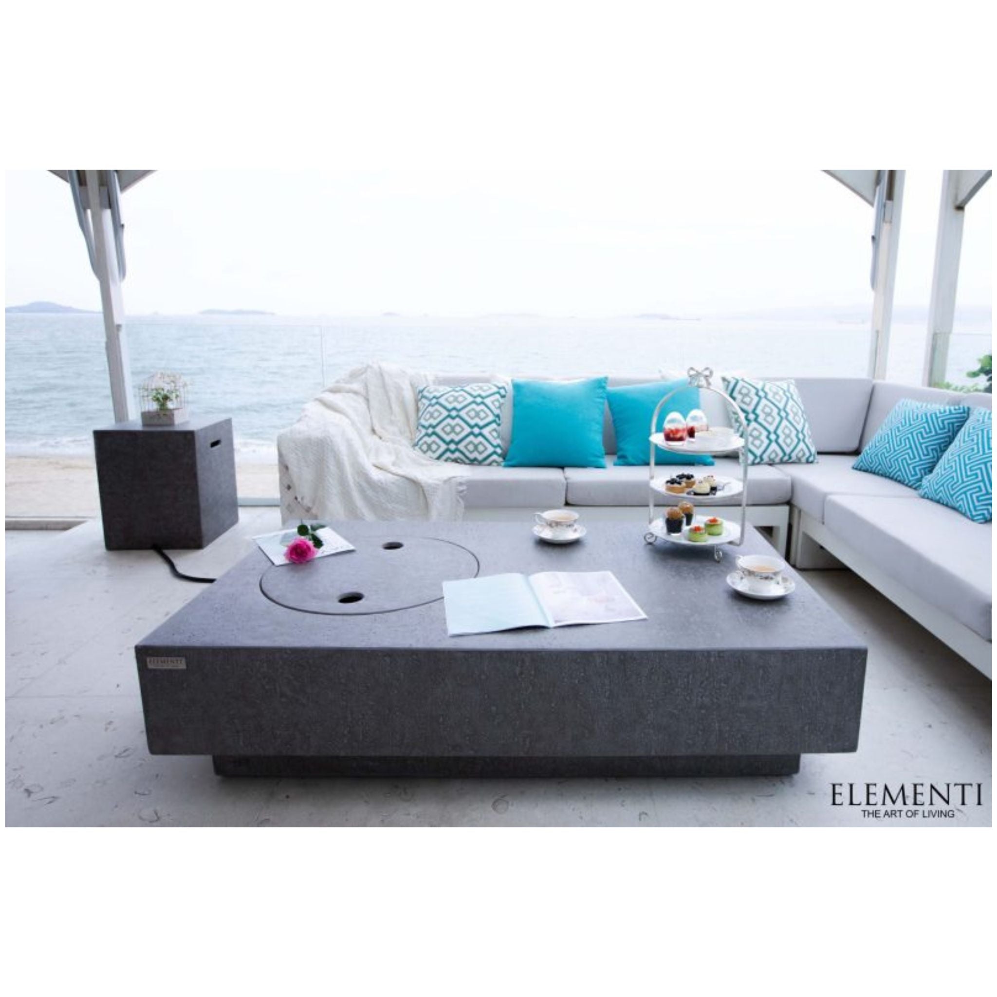 A modern outdoor patio scene featuring a large dark gray rectangular coffee table with a circular inset, set in front of a light gray sectional sofa with turquoise pillows. The scene is styled with a three-tiered tea stand, cups, and a small side table, with the ocean visible in the background.
