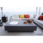 A large rectangular outdoor coffee table in dark gray stone or concrete, styled in a modern patio setting with a gray sectional sofa, colorful pillows, and a view of the sea in the background. The brand name ELEMENTI is visible in the bottom right corner.
