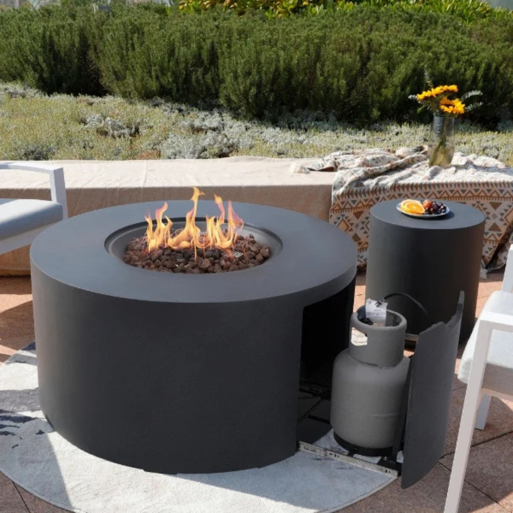 Round outdoor fire pit with flames, surrounded by greenery and outdoor furniture.