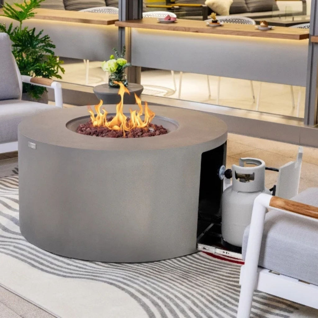 A round concrete fire pit table with a visible flame and fire rocks, shown in a modern living room setting next to a white chair and a propane tank.