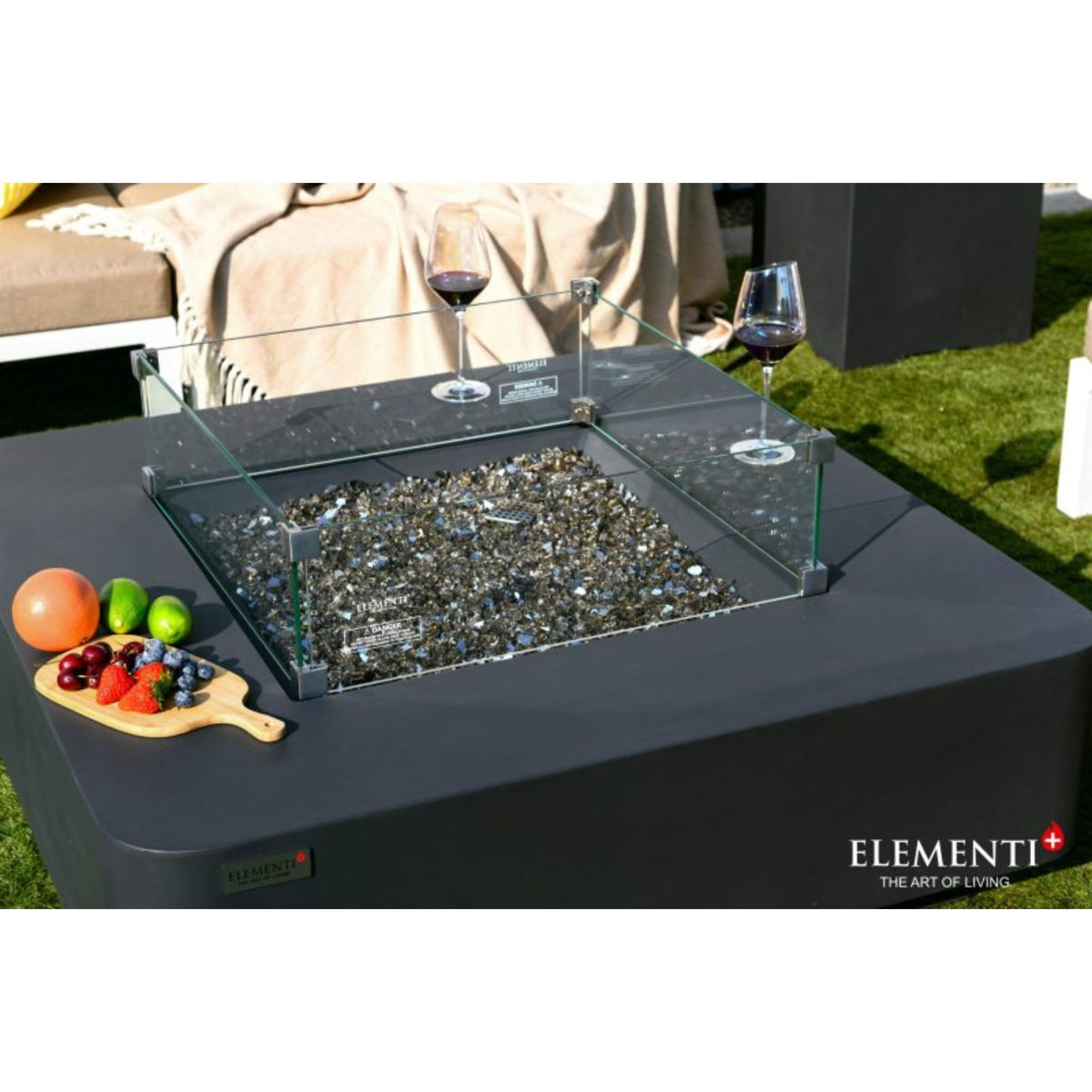 An outdoor Elementi fire pit table with a black square base, clear glass wind guard, and gray decorative stones inside. The table is set on grass next to a sofa, with wine glasses, fruit, and a cutting board on top.