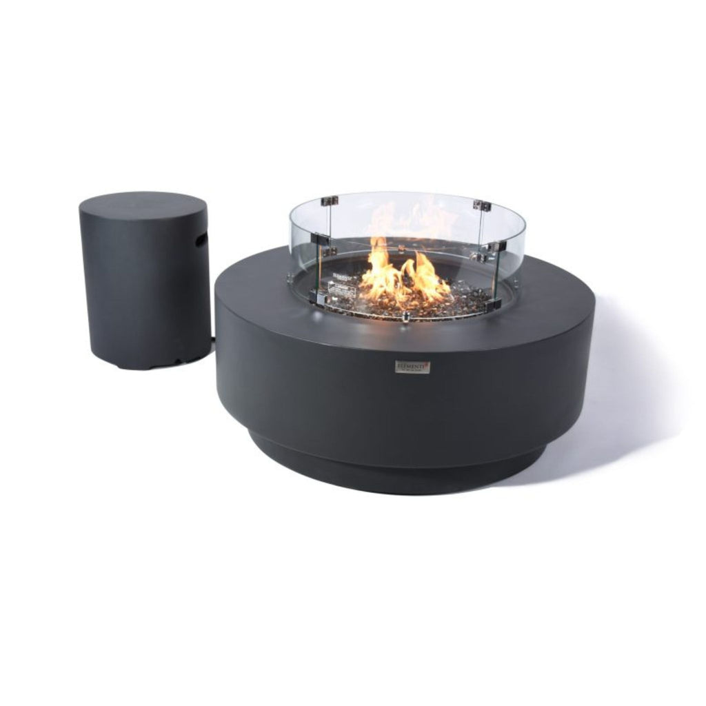 A round black outdoor fire pit table with a visible flame and clear glass wind guard, shown alongside a matching cylindrical black side table on a white background.