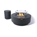 A round black outdoor fire pit table with a visible flame and clear glass wind guard, shown alongside a matching cylindrical black side table on a white background.