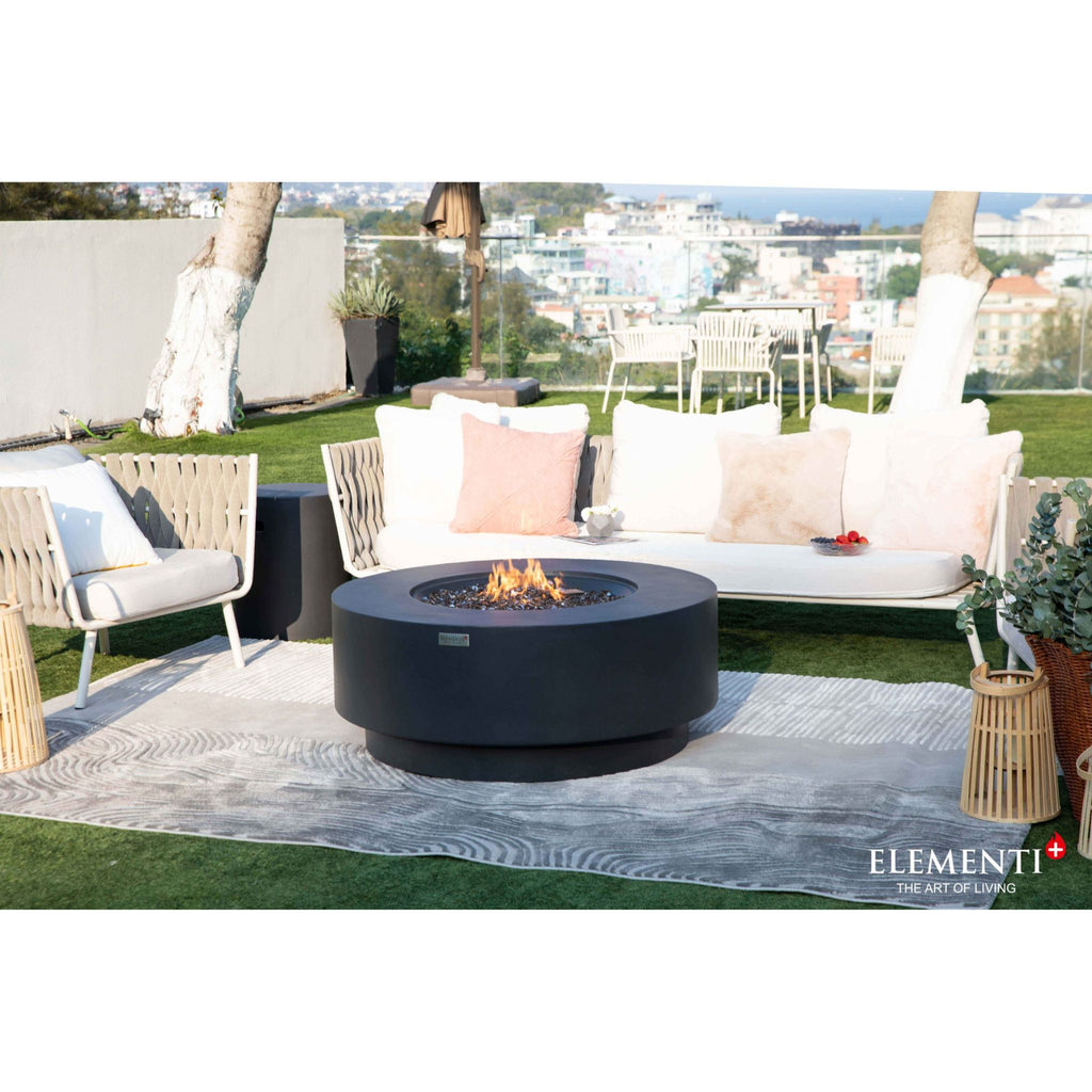 A round black outdoor fire pit with flames burning in the center, placed on a patterned gray rug in front of a white outdoor sofa with pink and beige pillows. The setting is a patio with a view of a coastal city, featuring an Elementi+ brand logo in the bottom right corner.