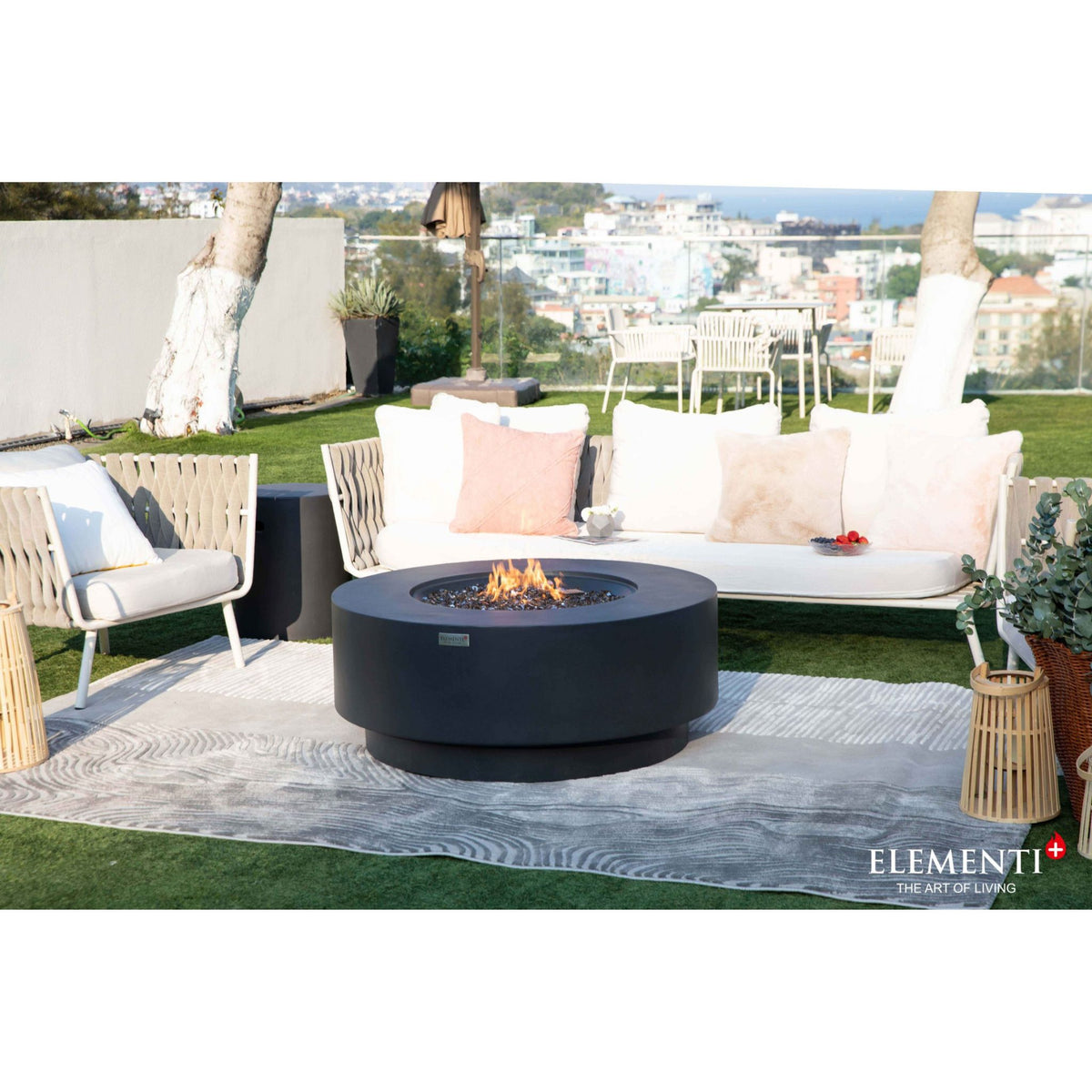 A round black outdoor fire pit with flames burning in the center, placed on a patterned gray rug in front of a white outdoor sofa with pink and beige pillows. The setting is a patio with a view of a coastal city, featuring an Elementi+ brand logo in the bottom right corner.