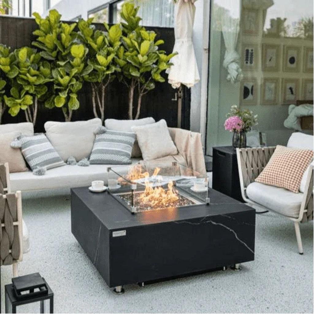 A modern outdoor patio scene featuring a black square fire pit table with a glass wind guard and visible flames, styled with a white sofa, striped pillows, a chair, and green plants in the background.