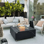 A modern outdoor patio scene featuring a black square fire pit table with a glass wind guard and visible flames, styled with a white sofa, striped pillows, a chair, and green plants in the background.