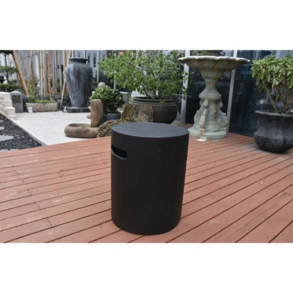A matte black cylindrical outdoor stool with a small side cutout, placed on a reddish-brown wooden deck in a garden setting with potted plants and stone garden furniture in the background.