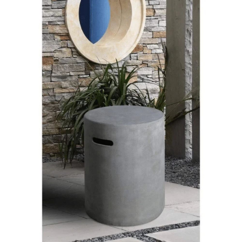 A cylindrical concrete storage bin or planter in a gray finish, featuring a cutout handle on the side, set outdoors against a stone wall with a potted plant behind it.