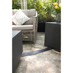 A black rubber floor drain with the Elementi brand name, installed on a stone patio next to a wicker sofa and dark planters.