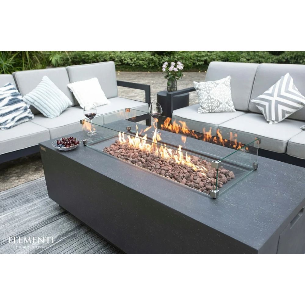 A modern outdoor patio scene featuring a rectangular dark gray fire pit table with a glass wind guard and active flames, surrounded by a matching gray sectional sofa with patterned throw pillows.
