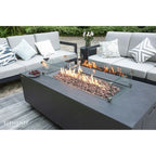 A modern outdoor patio scene featuring a rectangular dark gray fire pit table with a glass wind guard and active flames, surrounded by a matching gray sectional sofa with patterned throw pillows.