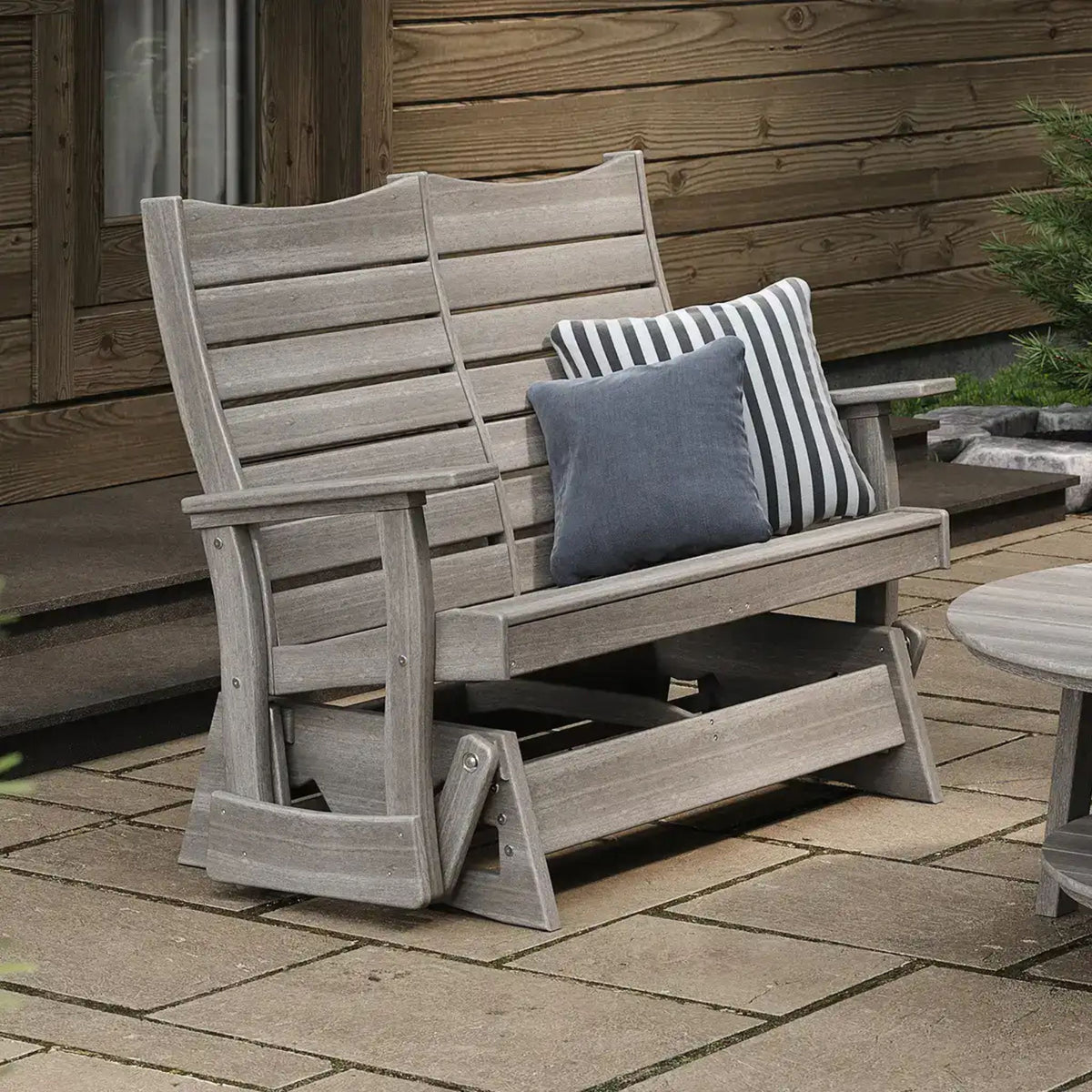 A weathered gray wooden outdoor glider chair with two throw pillows, one solid blue-gray and one black and white striped, set on a stone patio in front of a wooden house wall.