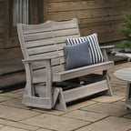 A weathered gray wooden outdoor glider chair with two throw pillows, one solid blue-gray and one black and white striped, set on a stone patio in front of a wooden house wall.