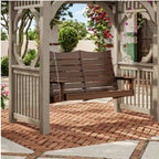 A two-person wooden porch swing hanging from chains within a light beige wooden pergola on a red brick patio, with greenery and flowers in the background.