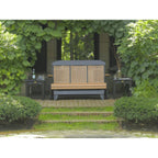 A wooden outdoor bench with a dark gray cushioned top and base, set on a paved patio in a garden courtyard with green foliage and ivy.