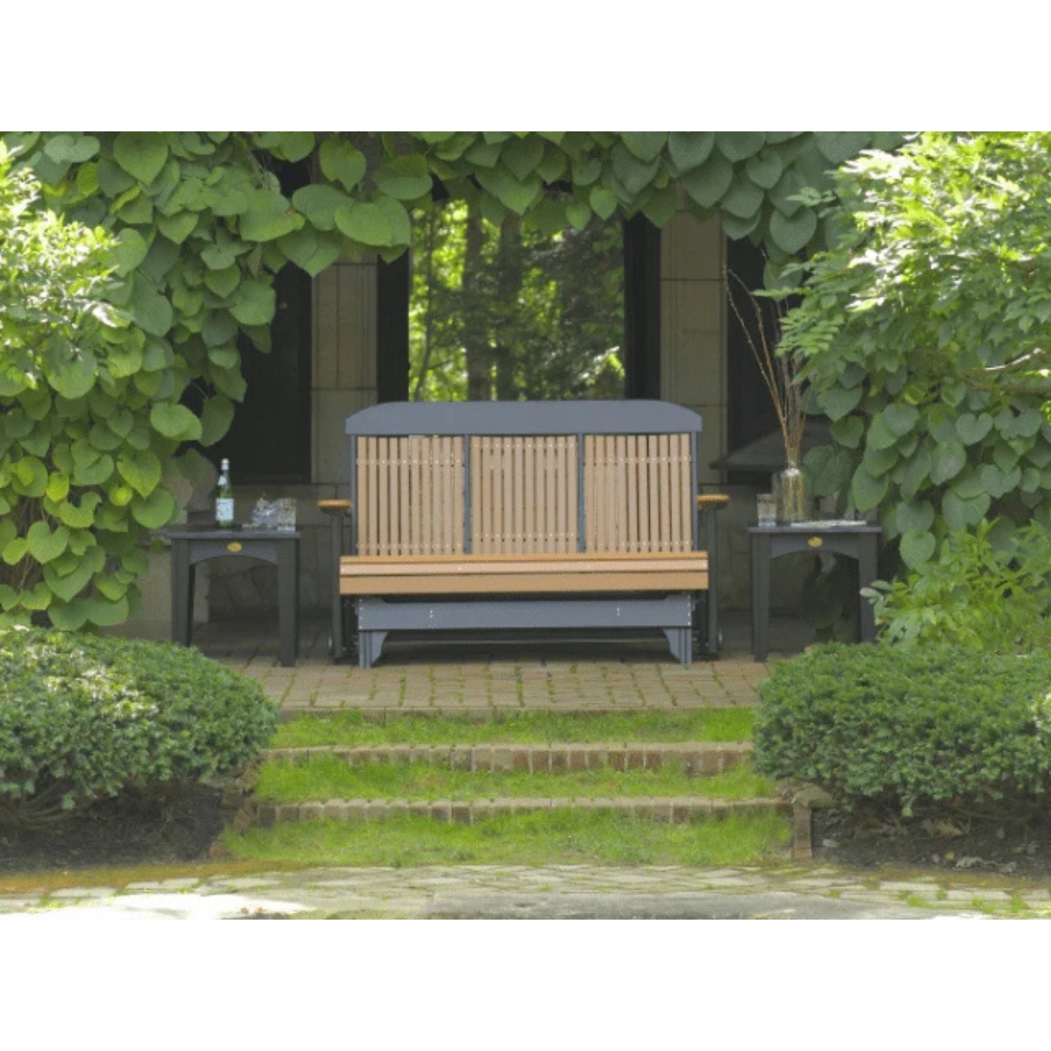 A wooden outdoor bench with a dark gray cushioned top and base, set on a paved patio in a garden courtyard with green foliage and ivy.