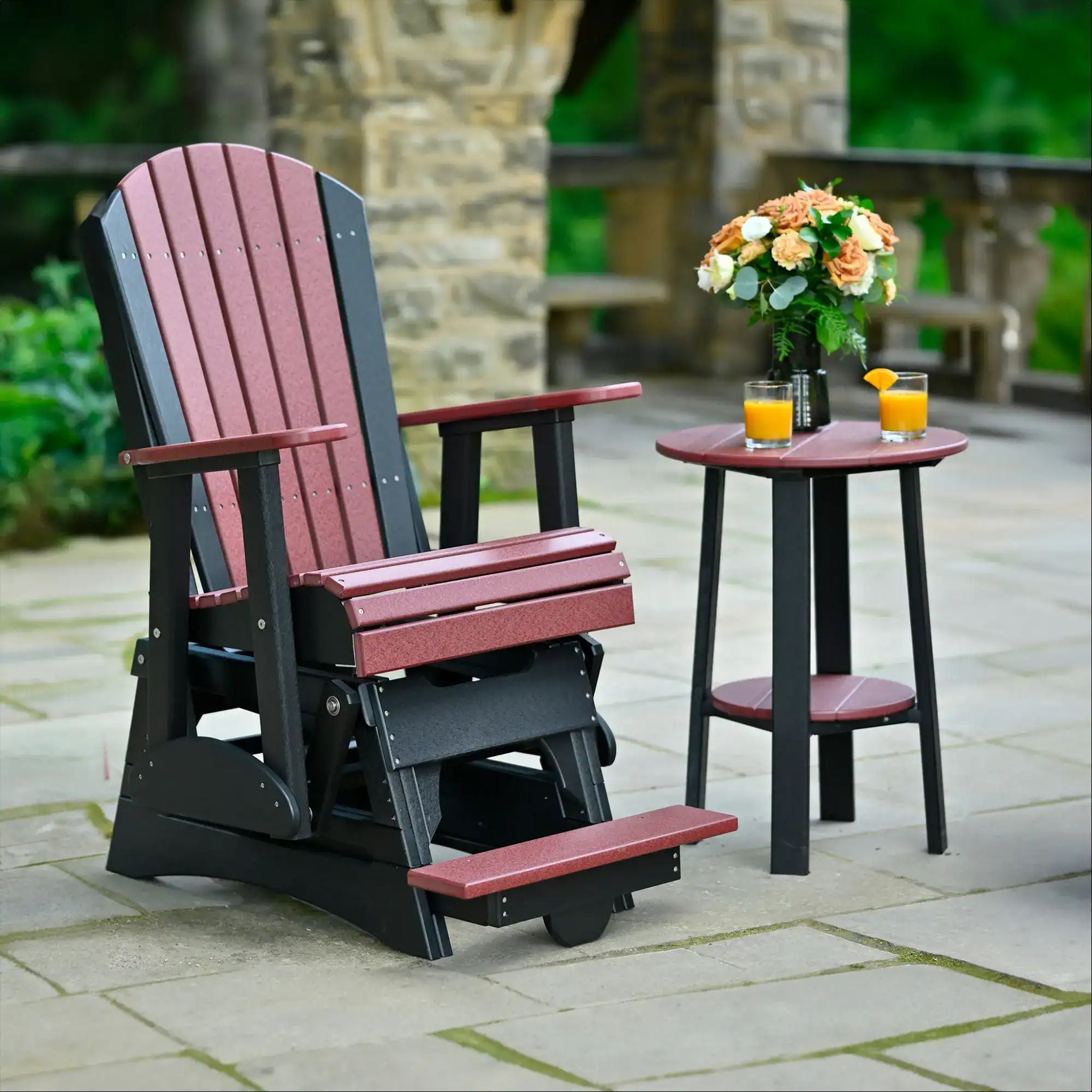 A dark red and black Adirondack-style outdoor glider chair with a matching footrest, shown beside a small dark red and black side table on a stone patio. The background features a stone column and green foliage.