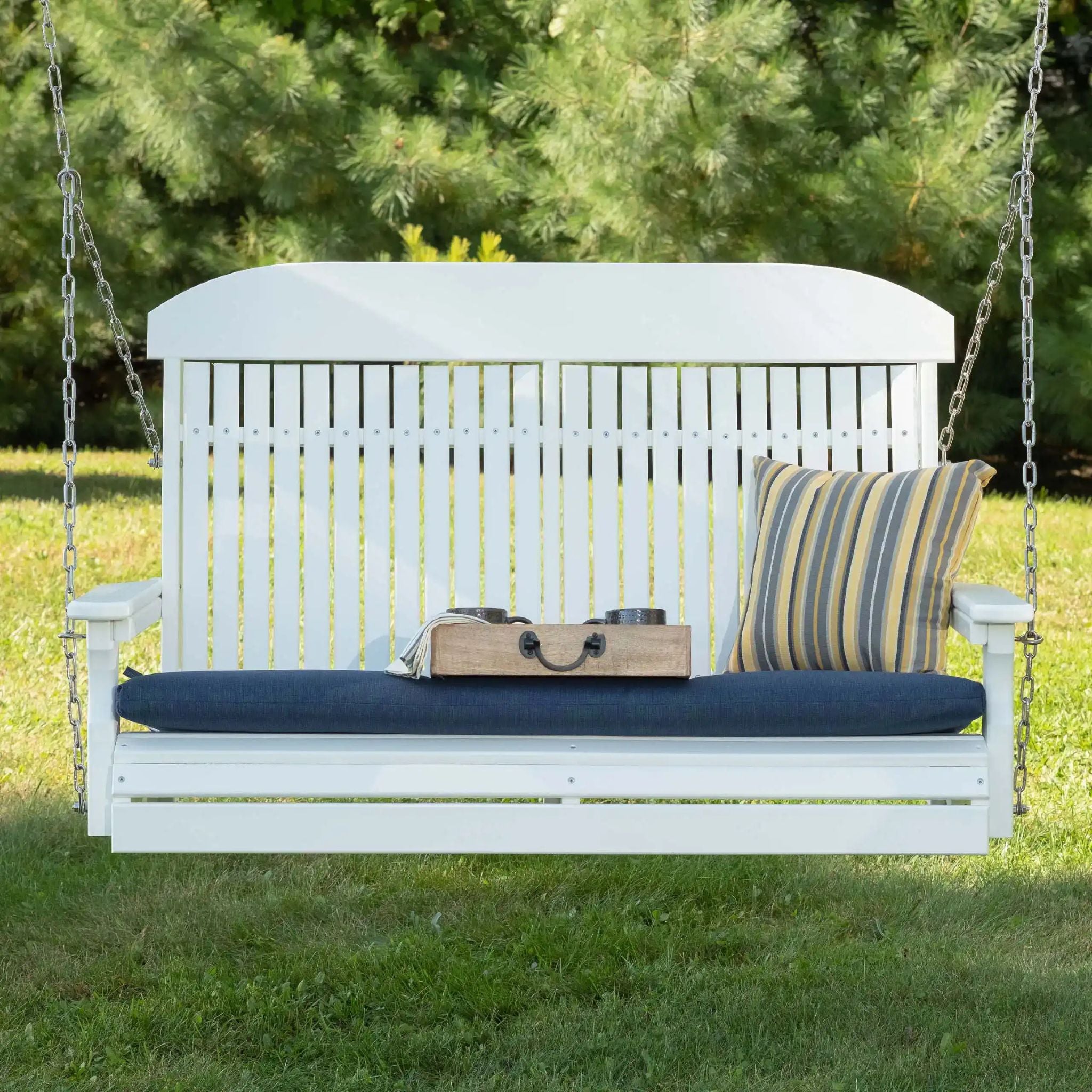 White wooden swing with blue cushion and striped pillow on a grassy area with trees in the background