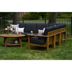 Outdoor wooden patio furniture set on a grassy lawn, including a curved sectional sofa with black cushions, a matching armchair, and a round wooden coffee table with a flower arrangement.