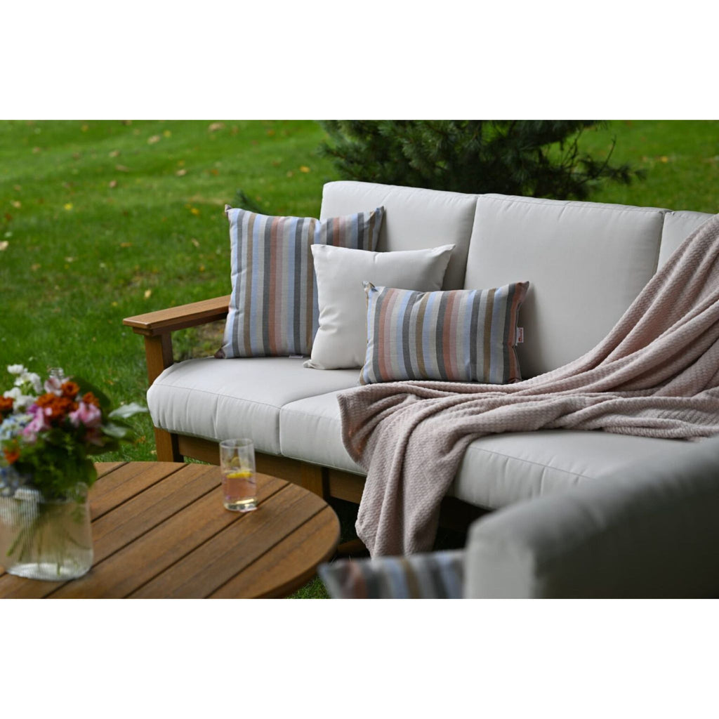 Outdoor patio seating set with light beige cushions on a wooden frame, styled with striped and solid accent pillows and a draped blush throw blanket, with a wooden table and green lawn in the background.