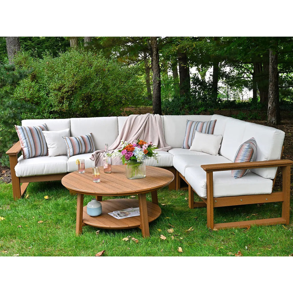 Outdoor sectional sofa set with white cushions and a round wooden coffee table on a green lawn, surrounded by trees. The set includes striped accent pillows and a draped throw blanket.