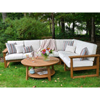 Outdoor sectional sofa set with white cushions and a round wooden coffee table on a green lawn, surrounded by trees. The set includes striped accent pillows and a draped throw blanket.