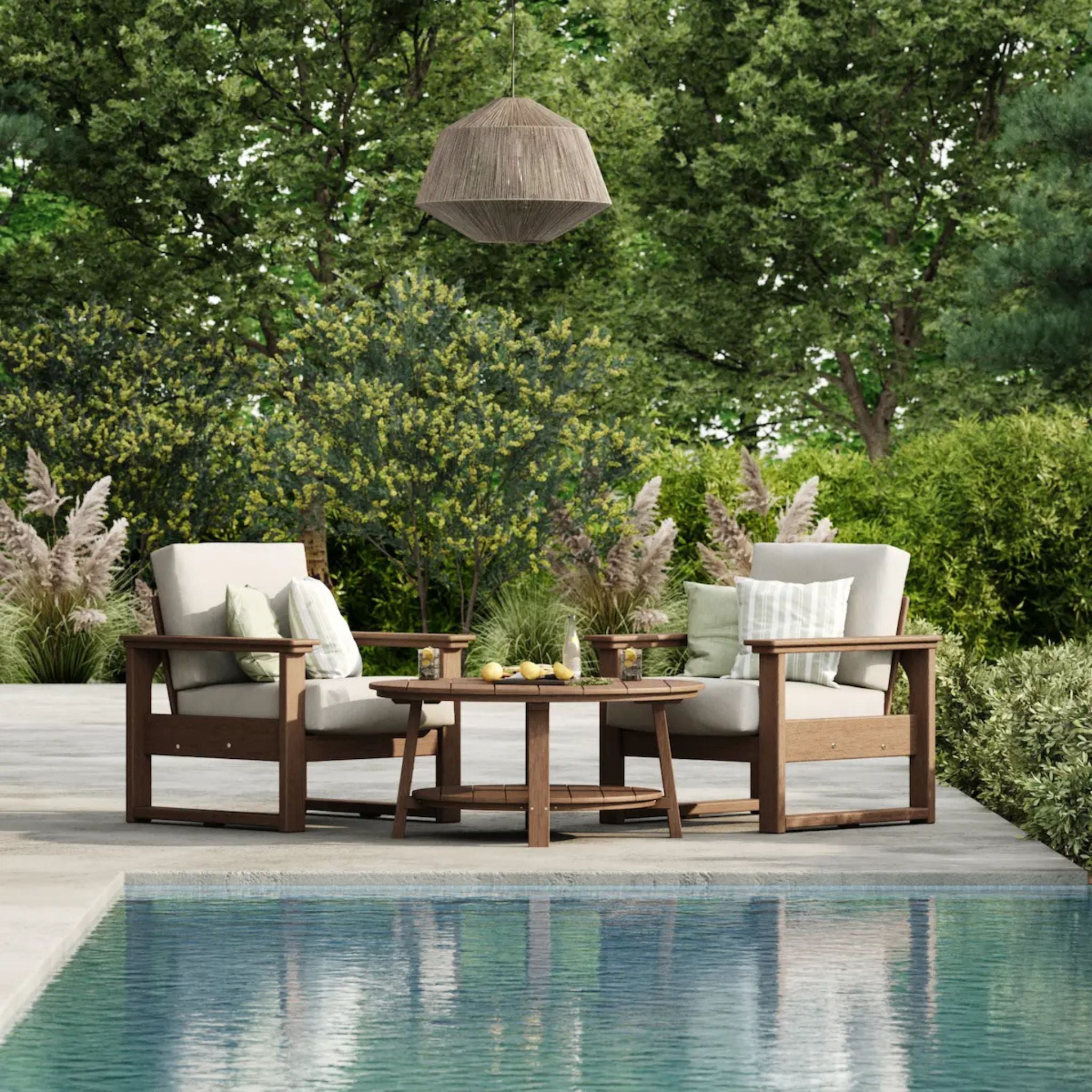 Two wooden outdoor armchairs with cream cushions and a matching round coffee table arranged on a patio beside a swimming pool, with lush green trees and shrubs in the background.