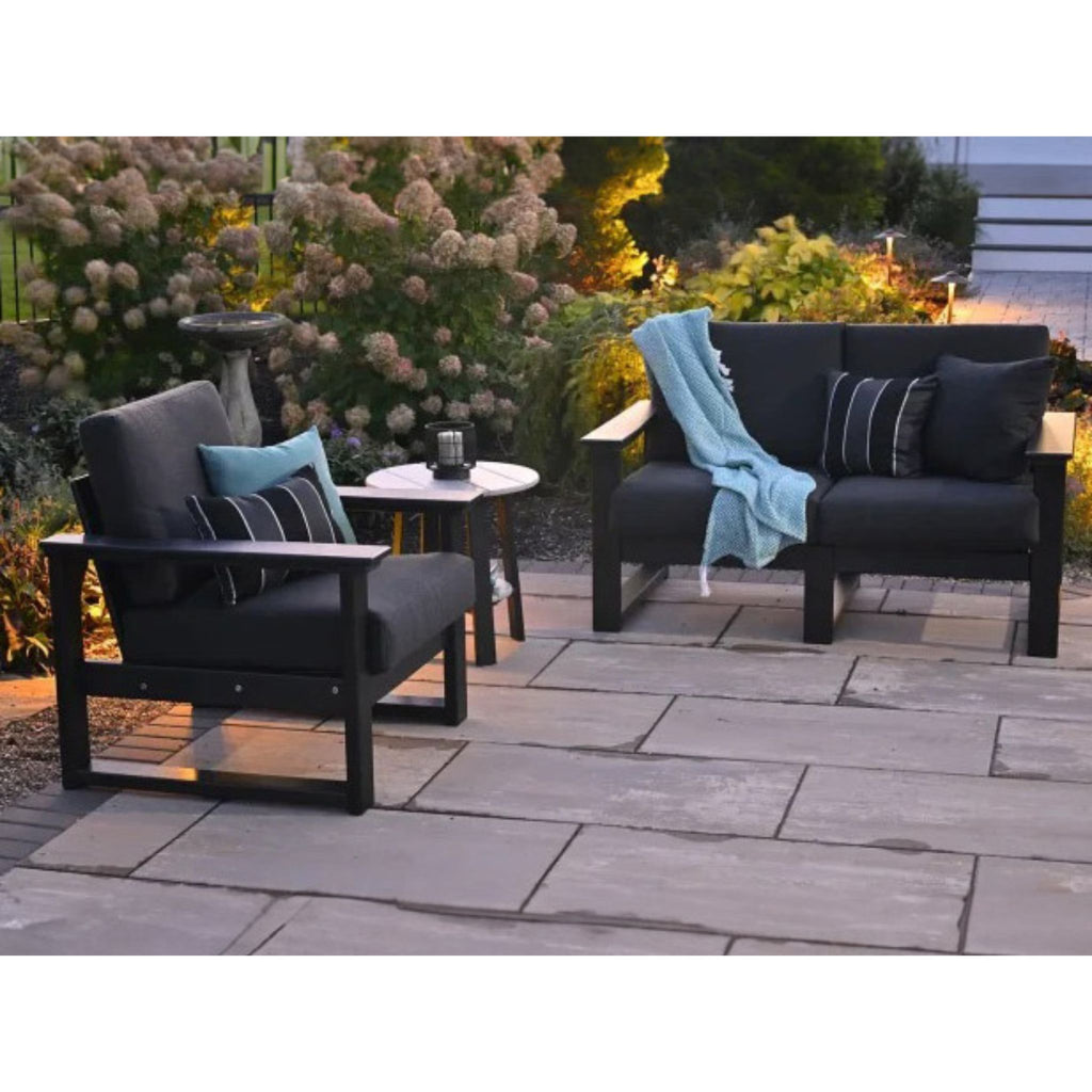 Outdoor patio furniture set including a black armchair, a black loveseat with a light blue throw blanket, and a small round side table on a stone patio with garden plants in the background.