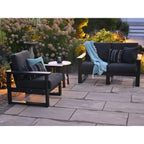 Outdoor patio furniture set including a black armchair, a black loveseat with a light blue throw blanket, and a small round side table on a stone patio with garden plants in the background.