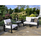 Outdoor patio furniture set including a black-framed armchair, loveseat, and small round side table with white cushions, styled on a stone patio in a landscaped garden.