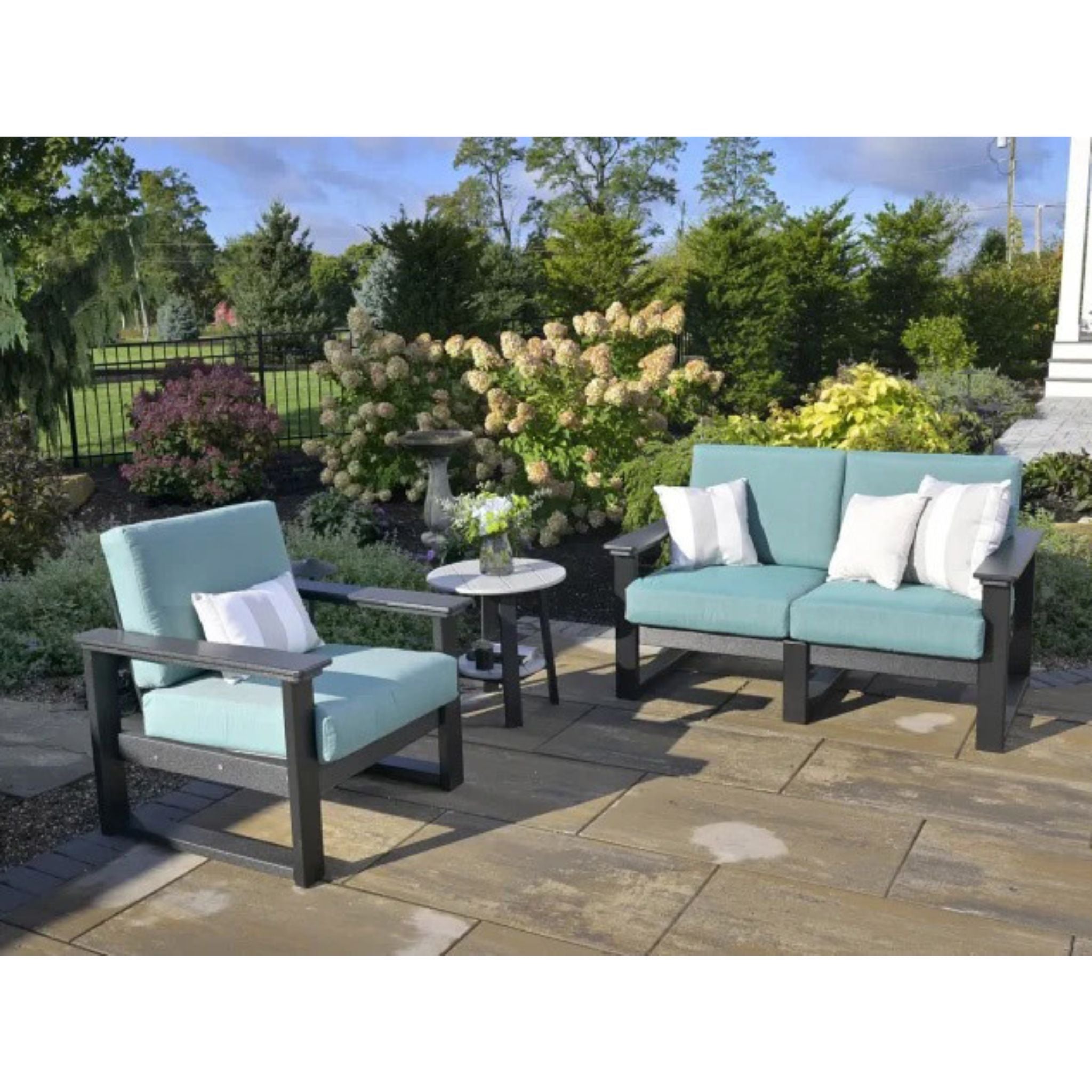 Outdoor patio furniture set including a two-seat sofa, a matching armchair, and a small round side table on a stone patio with a landscaped garden background.