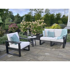 Three-piece outdoor patio furniture set including a loveseat, an armchair, and a small round side table, with white cushions, dark gray frames, and light blue accent pillows on a stone patio in a landscaped garden.