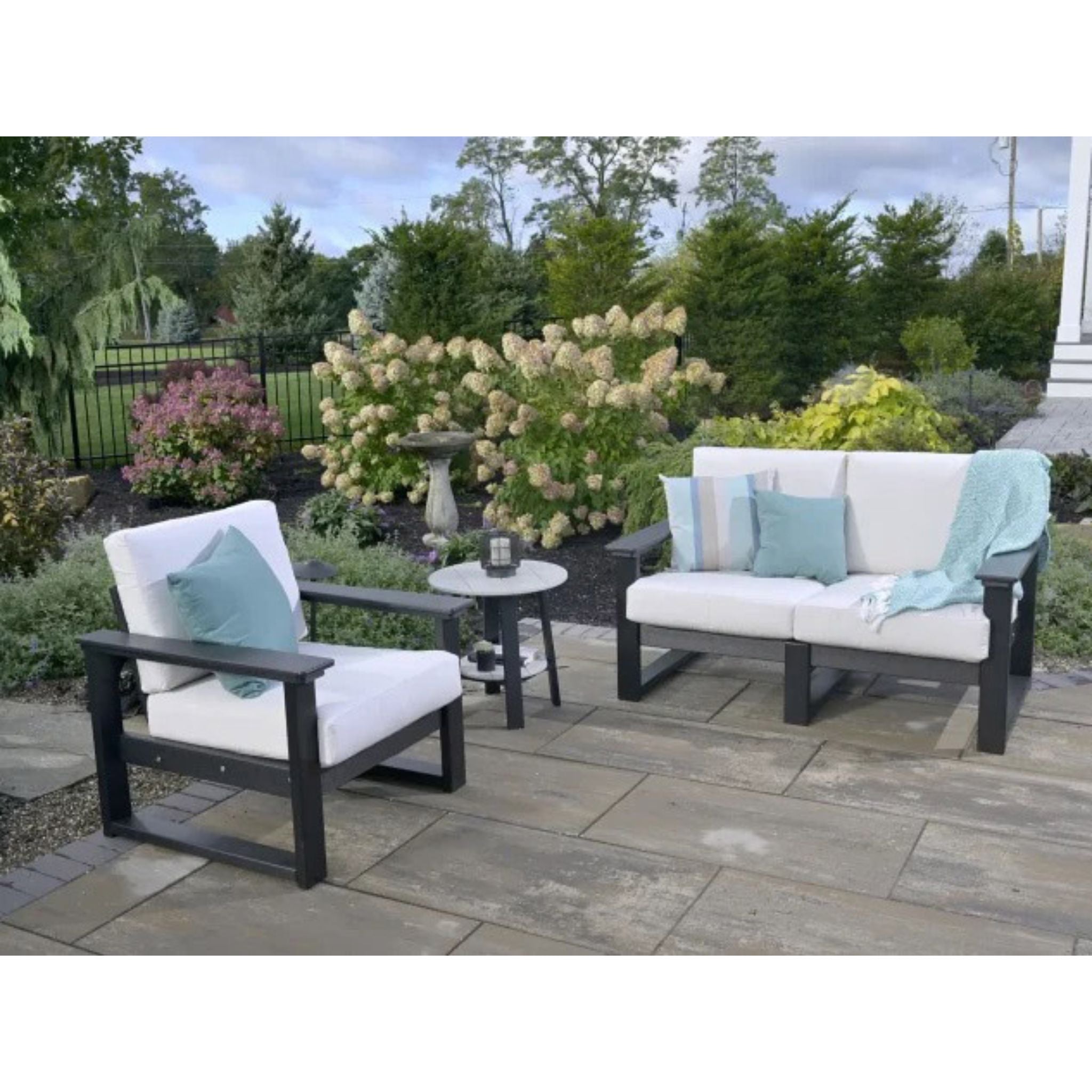 Three-piece outdoor patio furniture set including a loveseat, an armchair, and a small round side table, with white cushions, dark gray frames, and light blue accent pillows on a stone patio in a landscaped garden.