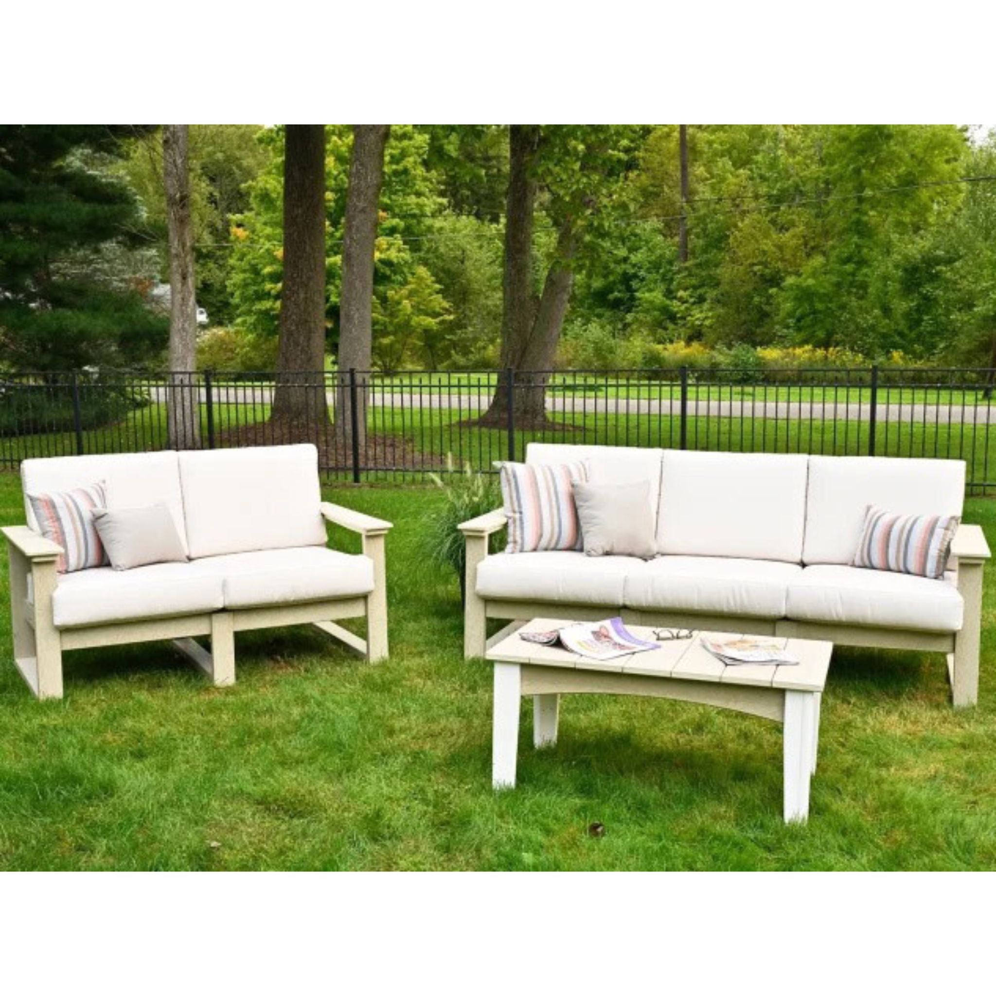 Outdoor patio furniture set on a grassy lawn with trees in the background, including a white loveseat, a matching white sofa, and a white coffee table with striped accent pillows.