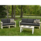 Outdoor patio furniture set on a grassy lawn with trees and a fence in the background, including a loveseat, a sofa, and a rectangular coffee table. The furniture has light natural wood frames and dark charcoal gray cushions, with two striped throw pillows.