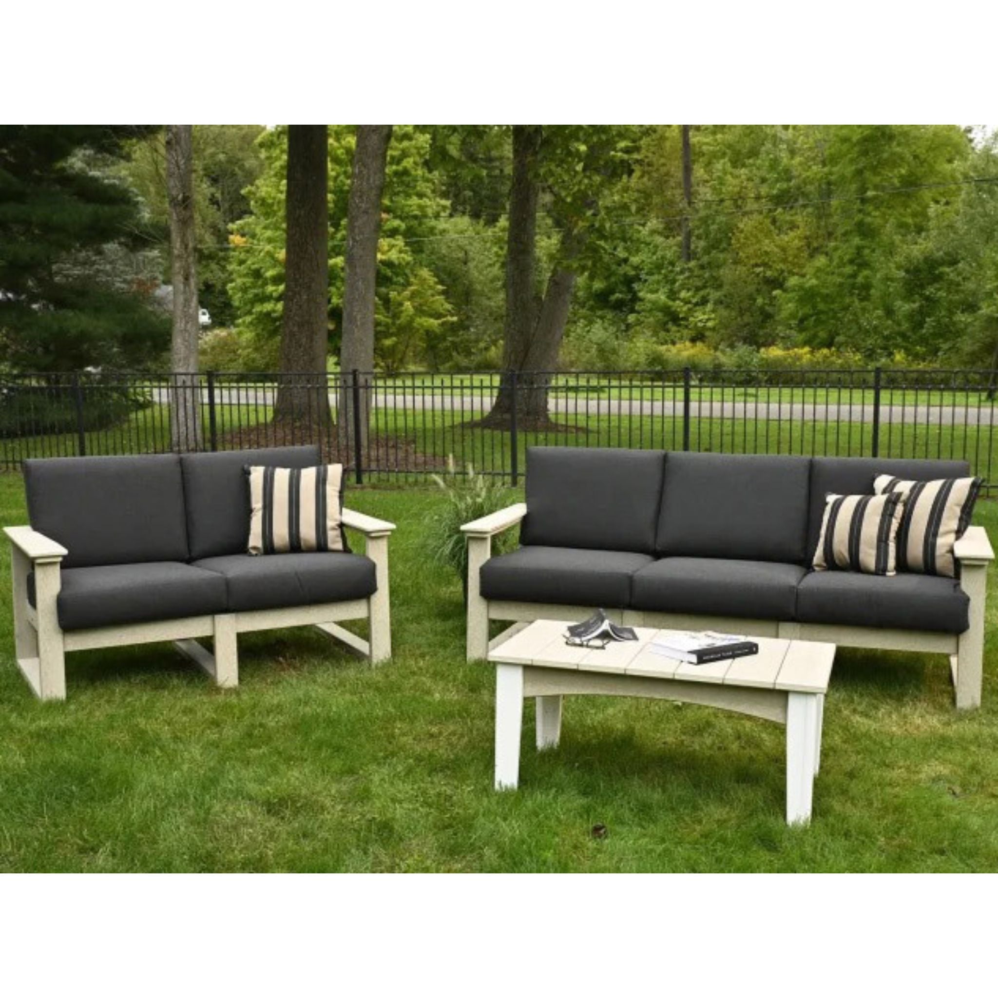 Outdoor patio furniture set on a grassy lawn with trees and a fence in the background, including a loveseat, a sofa, and a rectangular coffee table. The furniture has light natural wood frames and dark charcoal gray cushions, with two striped throw pillows.