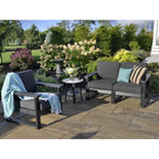 Three-piece outdoor patio furniture set including a loveseat, an armchair with a light blue throw blanket, and a small round side table, arranged on a stone patio in a landscaped garden setting.