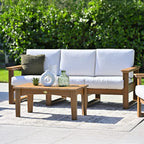 Outdoor patio set with white cushions, wooden coffee table, and greenery in the background.