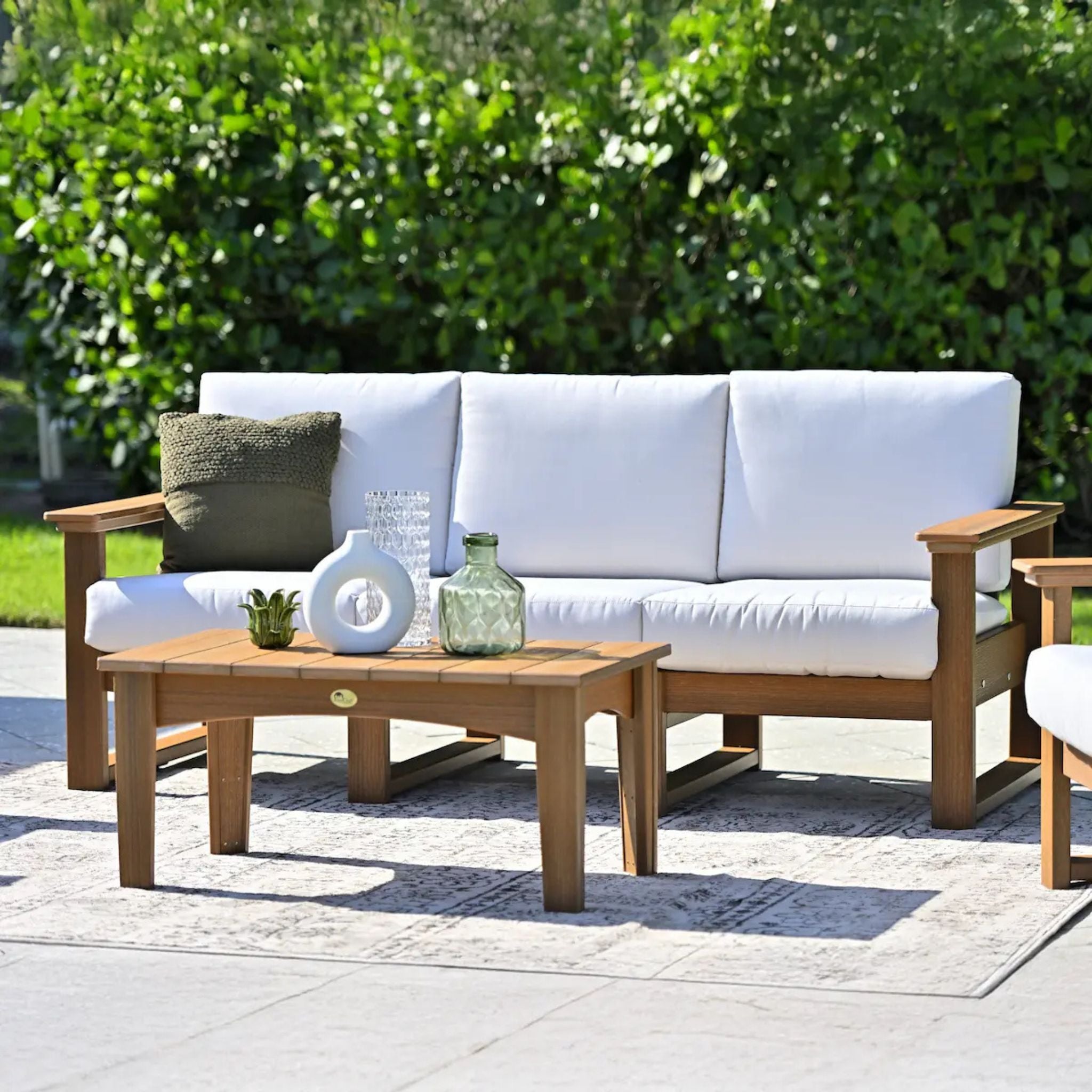 Outdoor patio set with white cushions, wooden coffee table, and greenery in the background.