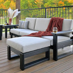 Modern outdoor sectional sofa set with a chaise ottoman, upholstered in light beige cushions and dark charcoal frames, styled on a wooden deck with a red throw blanket and a side table.