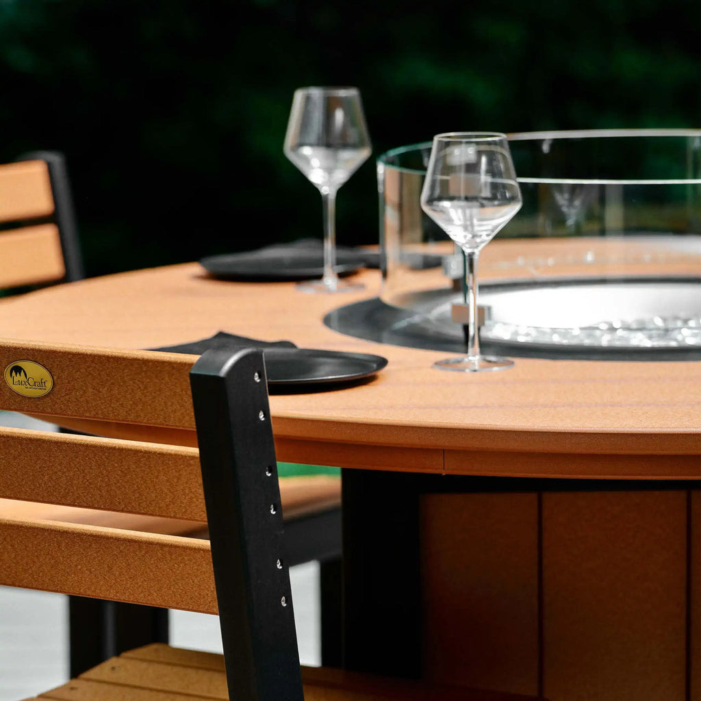 Outdoor round fire pit table with a warm brown wood-look finish, clear glass wind guard, black burner base, and matching wooden dining chairs with black metal frames.