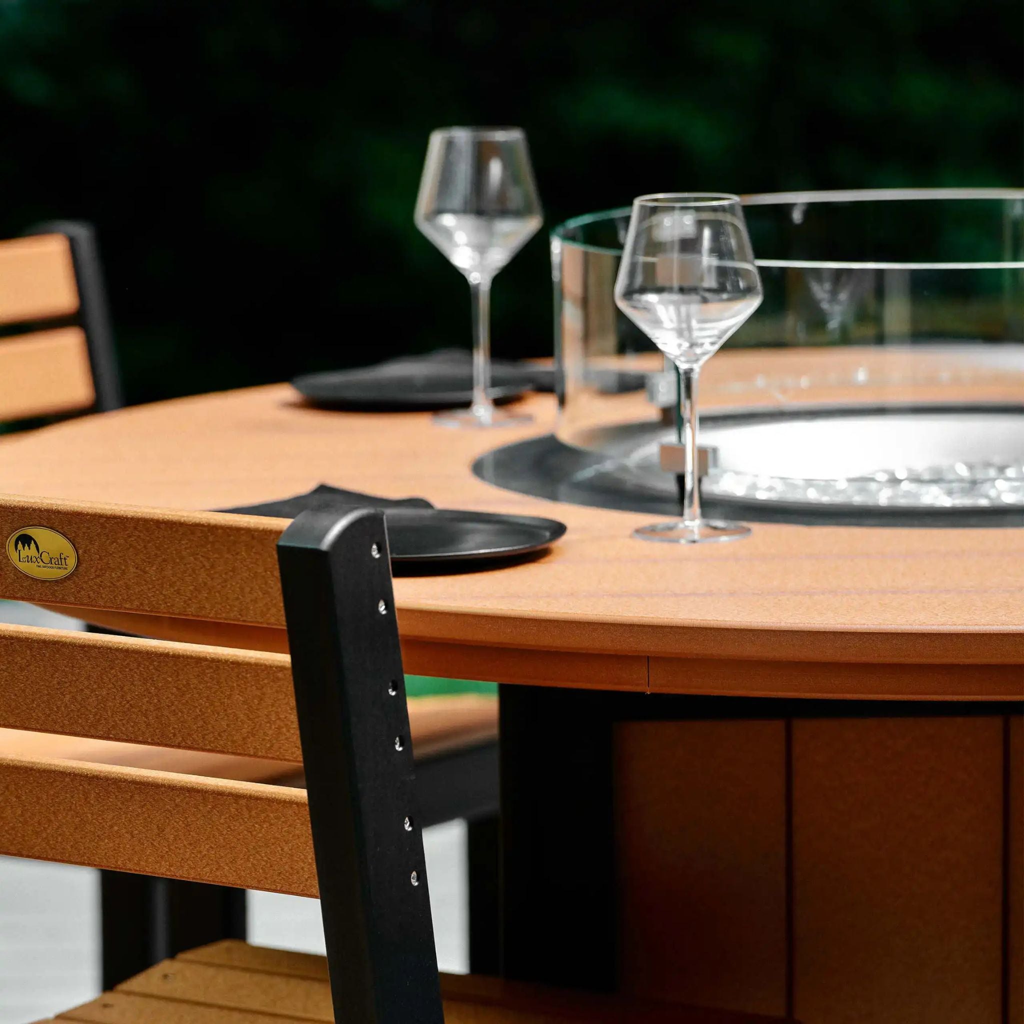 Outdoor round fire pit table with a warm brown wood-look finish, clear glass wind guard, black burner base, and matching wooden dining chairs with black metal frames.