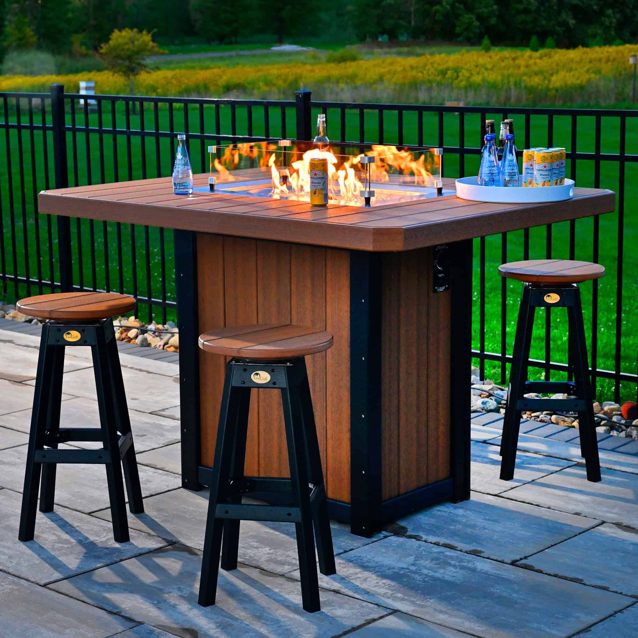Outdoor patio table with fire pit, stools, and drinks on a deck with greenery in the background