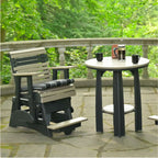 A dark gray and beige outdoor patio glider chair sits next to a matching round bar table on a stone patio with a lush green forest background.