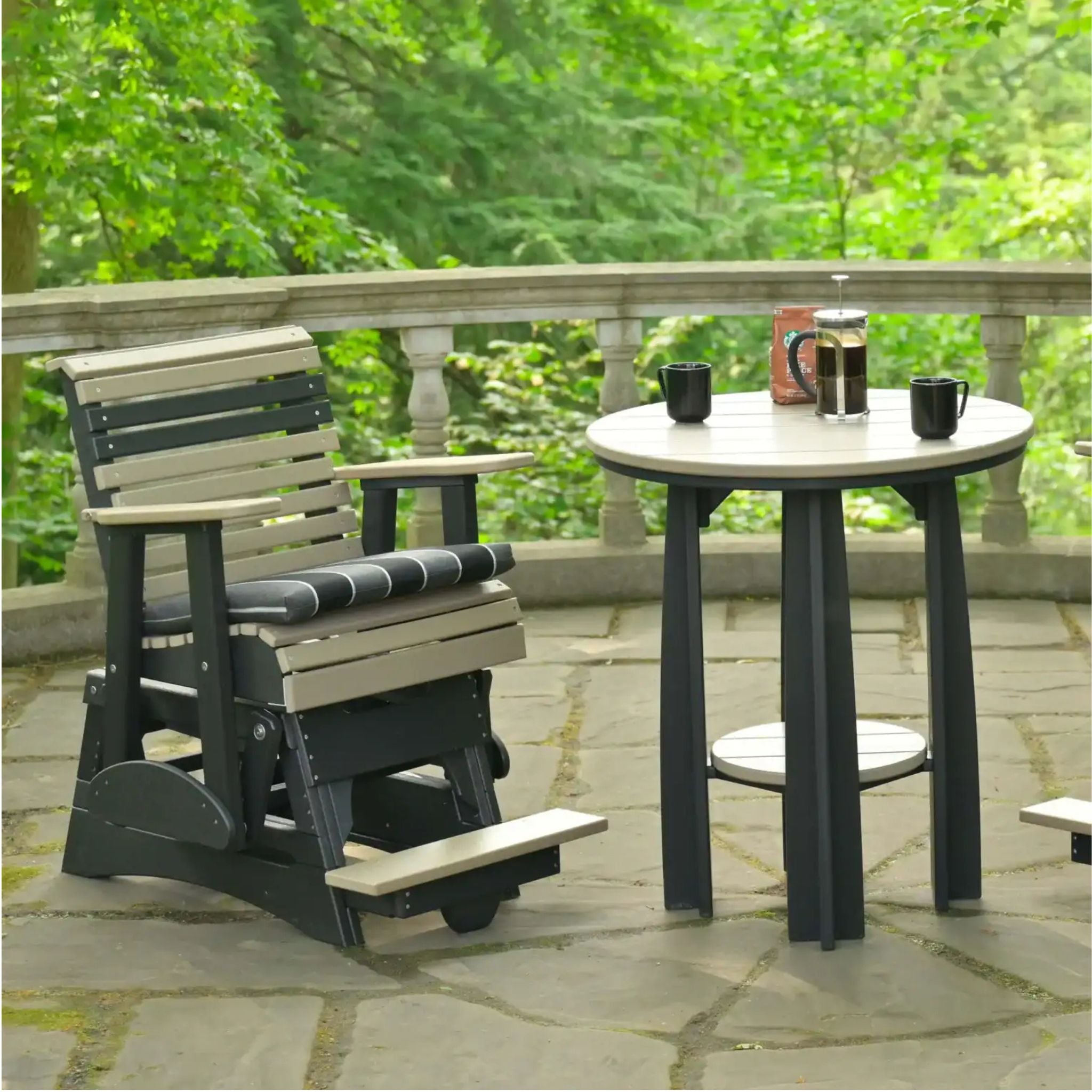A dark gray and beige outdoor patio glider chair sits next to a matching round bar table on a stone patio with a lush green forest background.