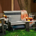 A gray and black outdoor patio bench with a slatted back and armrests, styled with an orange-and-white throw blanket, two iced tea glasses with lemon slices, and a glass pitcher on a small matching side table. The bench is set on grass in front of a wooden fence and tree trunk.